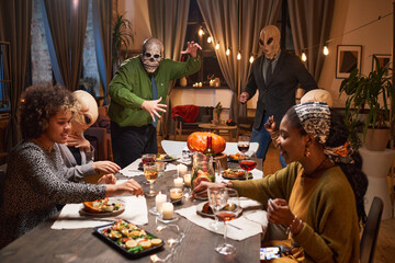 Group of people in scary costumes sitting at dining table and celebrating Halloween holiday at home