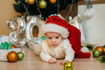 Cute smiling baby is lying under a festive Christmas tree and playing with gifts. Christmas and New Year celebrations