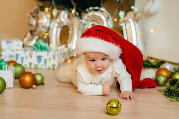 Cute smiling baby is lying under a festive Christmas tree and playing with gifts. Christmas and New Year celebrations