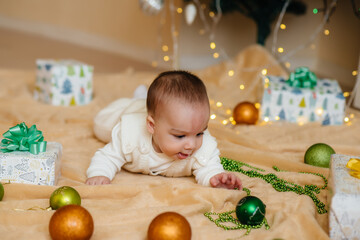Fototapeta premium Cute smiling baby is lying under a festive Christmas tree and playing with gifts. Christmas and New Year celebrations