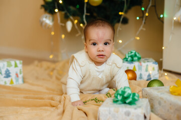 Cute smiling baby is lying under a festive Christmas tree and playing with gifts. Christmas and New Year celebrations