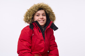 Waist length portrait of handsome cheerful preadolescent, school aged boy wearing bright red down jacket with hood, smiling toothy smile looking at camera isolated on white background with copy space © Taras Grebinets