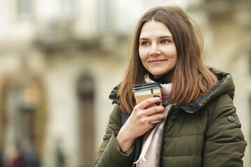 Coffee lover concept: smiling young woman drinking hot beverage at street of European city. Model wearing green coat, white and beige scarf. Close up. Outdoor shot