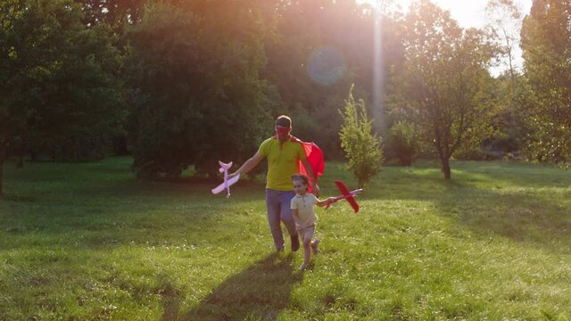 Happy and excited dad with his small son walking through the grass with a red airplane dad wearing superhero suit