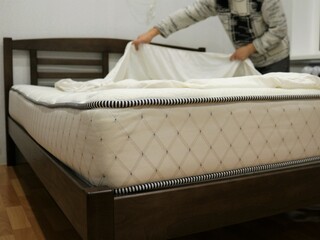 a man tucking a mattress topper on a double bed, filling a protective cover on a mattress of a large bed in a bedroom with a person in a blurry background, a man making a bed - obrazy, fototapety, plakaty
