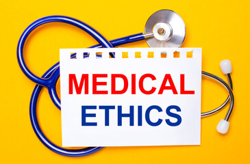 Fototapeta premium On a bright yellow background, a blue stethoscope and a sheet of paper with the text MEDICAL ETHICS. Medical concept