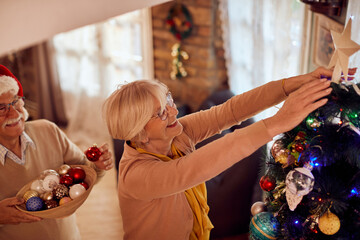 Happy senior woman putts star on top of Christmas tree while decorating home with her husband.