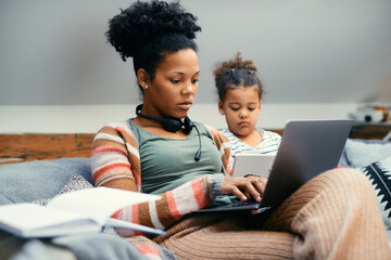African American working mother uses laptop while her daughter is using touchpad at home.