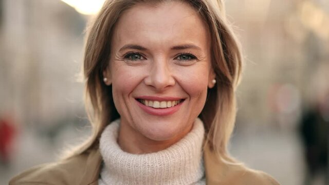 Lovely middle-aged blond woman with a beaming smile and looking at the camera. Happy adult woman smiling with teeth smile outdoors and stay on city street at sunset time