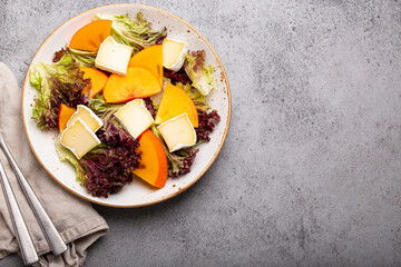 Top view flat lay persimmon salad with brie cheese, fresh salad leaves on white plate stone gray background, seasonal fruit salad as appetizer, vegetarian healthy food space for text
