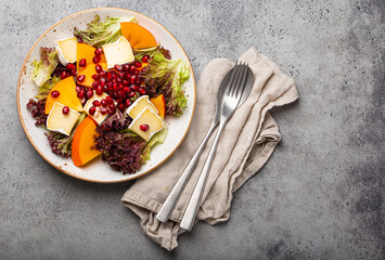 Top view flat lay persimmon salad with brie cheese, fresh salad leaves, pomegranate seeds on white plate stone gray background, seasonal fruit salad as appetizer, vegetarian healthy food copy space