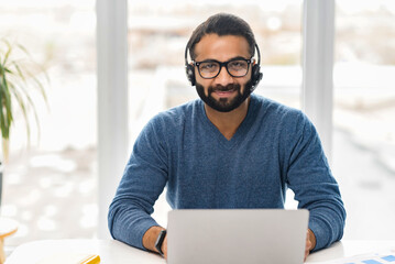 Indian male salesman in a headset is holding the call, ethnic customer service representative sitting in front of a laptop and looking at the camera