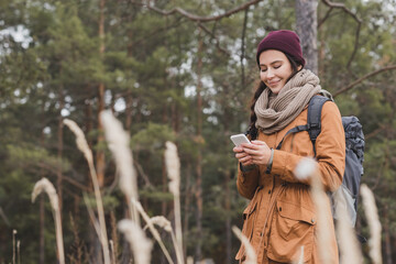 happy woman in autumn outfit using mobile phone during walk in forest