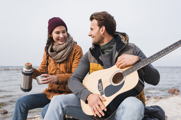 cheerful woman with thermos and man with guitar sitting at riverside during autumn walk