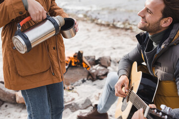 happy man playing guitar near woman pouring drink from thermos