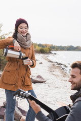 Naklejka premium cheerful woman pouring drink from thermos near blurred man playing guitar on sea coast