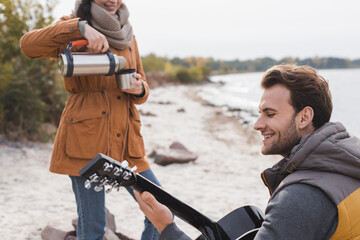 cheerful man playing acoustic guitar while woman pouring drink from thermos during autumn walk