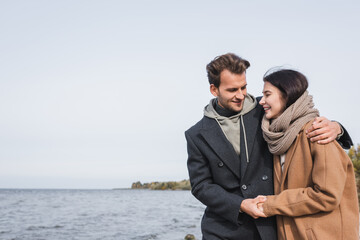 pleased couple in autumn clothes holding hands while walking near river