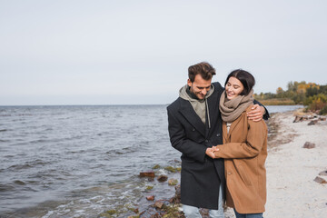 joyful couple in autumn coats hugging ad holding hands while walking along riverside