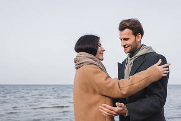 happy young couple in autumn coats looking at each other near sea