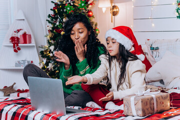 African American woman and brunette daughter in Santa hat sit among Christmas decorations and blow kisses chatting on video call via laptop - Powered by Adobe