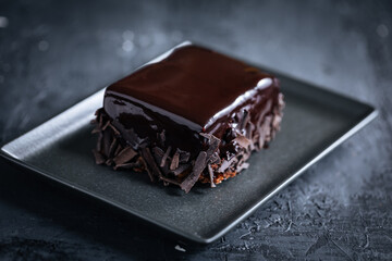 Glazed chocolate cake, one piece, on a dark black background on black plate with golden cake