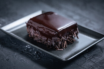 Glazed chocolate cake, one piece, on a dark black background on black plate with golden cake