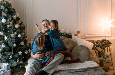 Dad and little daughters have fun together, sitting on the bed surrounded by pillows near the Christmas tree. happy christmas
