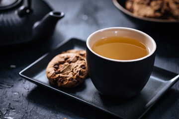 Dark grey cup of tea with cookies and teapot in background in a dark moody atmosphere 