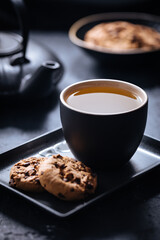 Dark grey cup of tea with cookies and teapot in background in a dark moody atmosphere 