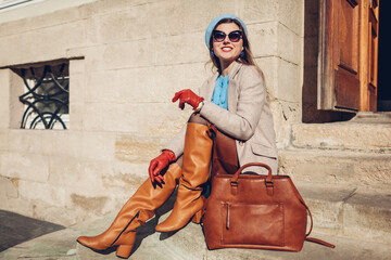 Portrait of woman wearing stylish orange boots coat beret sitting on stairs by handbag outdoors....