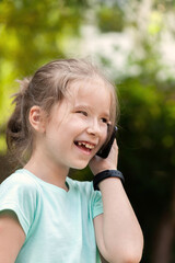 Happy cheerful elementary school age child, girl talking on her mobile phone, holding a smartphone next to ear wearing a smartwatch, laughing. Outdoors portrait, face closeup, communication concept