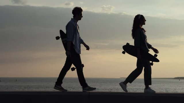 Couple of skateboarders walking on seaside carrying longboards at sunset. Silhouettes of two trendy skateboarders wear stylish urban style clothes enjoy sea view. Hipster lifestyle and freedom concept