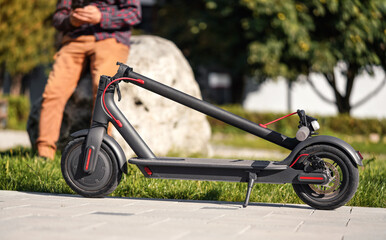 Folded electric scooter on concrete pavement, young man legs in background