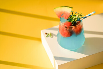 Blue margarita alcoholic cocktail with tequila on pedestal, liqueur,watermelon and ice, summer yellow background, copy space, shadows.