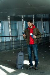 Fototapeta na wymiar Vertical portrait view of the attractive unshaven man standing at the airport lounge with telephone and smiling while checking something at the app