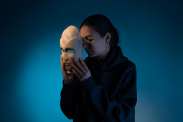 Hiding behind a mask, a young woman in a dark hoodie hides her face with a mask, self-identification problems and impostor syndrome. Portrait in the studio on a dark gray background.