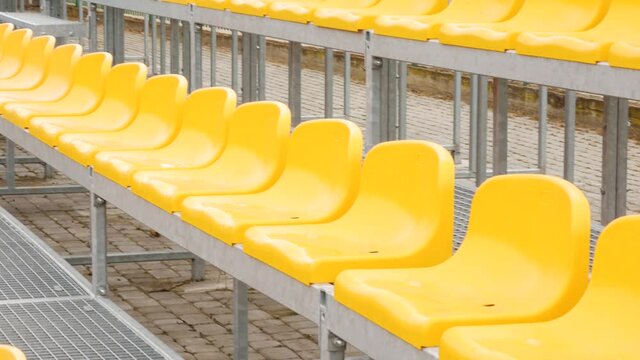 Vacant empty arena stadium stands, chairs, plastic yellow spectator seats on a small soccer football field, daytime, pan, nobody. No people at a sport event, fans absence, rows of seats outdoors