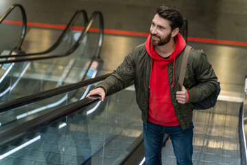 Nice unshaven man wearing backpack standing at the moving staircase while going at the airport and...