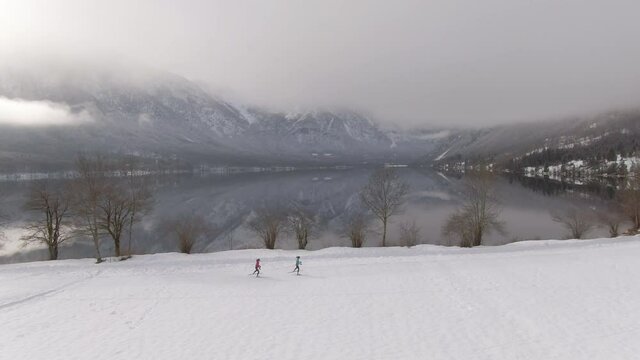 AERIAL: Two female athletes Nordic ski around the breathtaking lake Bohinj on a foggy winter day. Spectacular drone shot of Nordic skiers training in the picturesque snow covered Slovenian mountains.
