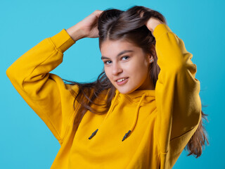 Fototapeta na wymiar A beautiful young brunette in a yellow oversize hoodie, a positive student in a bright outfit, portrait on a blue background