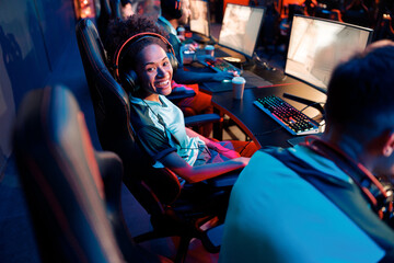 Fototapeta na wymiar African female cybersport gamer wearing headset sitting on gaming chair and looking at camera with cheerful expression in cyber club