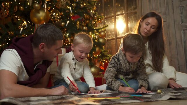 Young parents with two young sons draw sitting on the floor. The family celebrates, spends time together on Christmas Eve or New Year's Eve