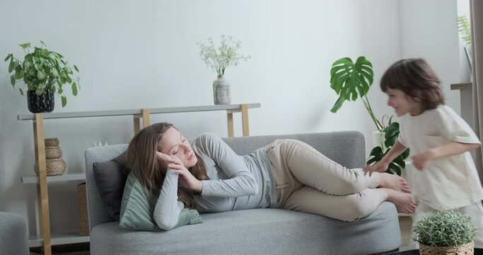 Tired woman lying on couch relaxing at cosiness home interior. Cute little boy kid running having fun near mother enjoying happy childhood. Family resting have time for yourself weekend recreation