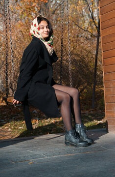 A beautiful girl sits in the park on a swing, on her head is a traditional Russian headscarf