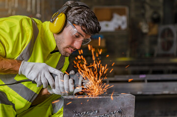 In a factory, a young man is grinding steel structures with an electric wheel.