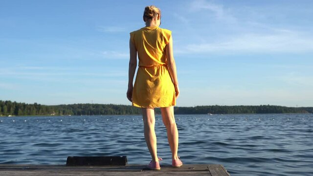 Blonde girl on shore of Baltic sea bay. Young woman in yellow dress walks on the pier and looks into the distance at sea. Panorama of the Stockholm archipelago with forest islands. Summer day on coast