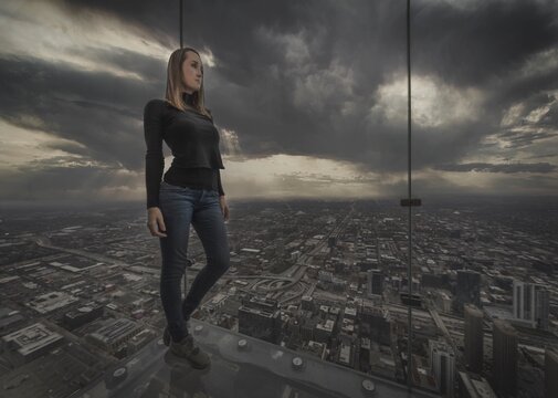 This image showcases a young woman standing in a glass environment with an aerial view , looking out over the city of Chicago on a dramatic weather day.