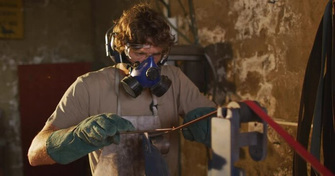 Caucasian male blacksmith wearing breathing mask and ear guards, forging metal tool in workshop