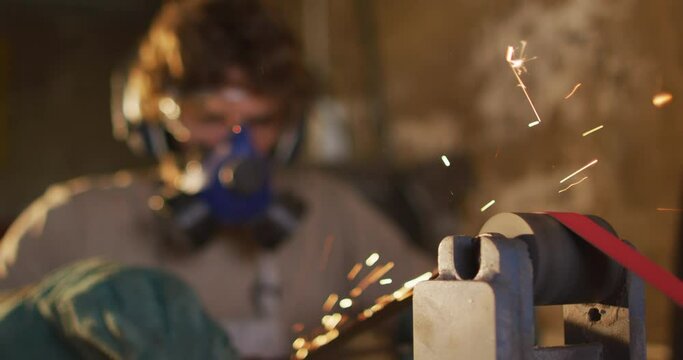 Caucasian male blacksmith wearing breathing mask and ear guards, forging metal tool in workshop